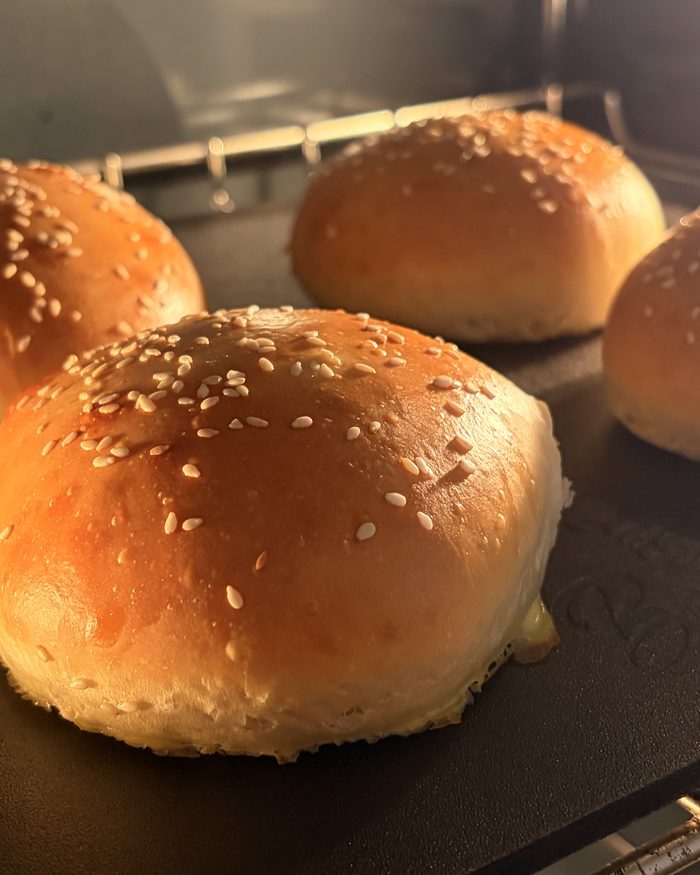 Four golden sesame-topped hamburger buns baking on a tray.